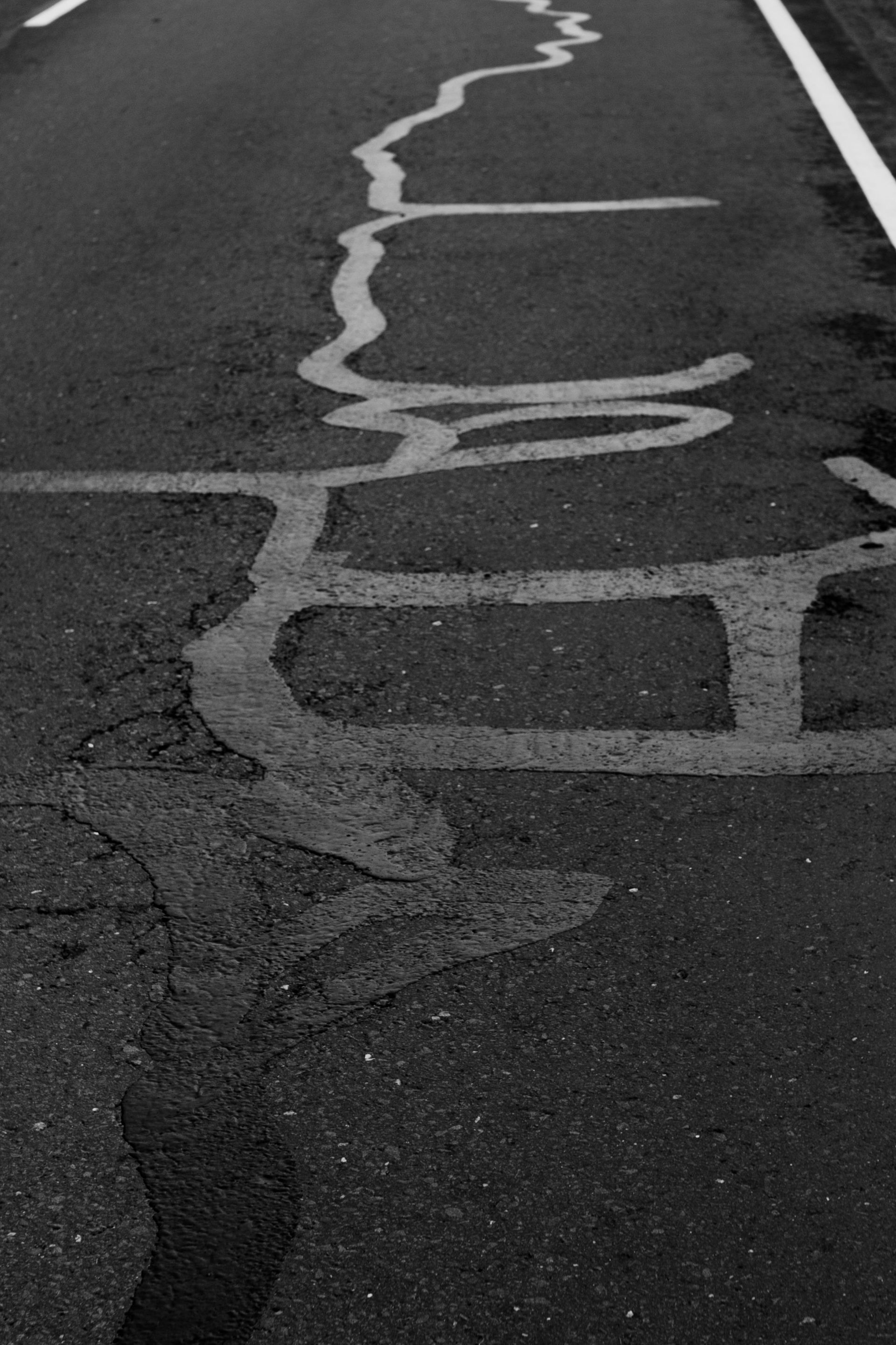 A detailed close-up of a cracked asphalt road with visible repair patches in grayscale.